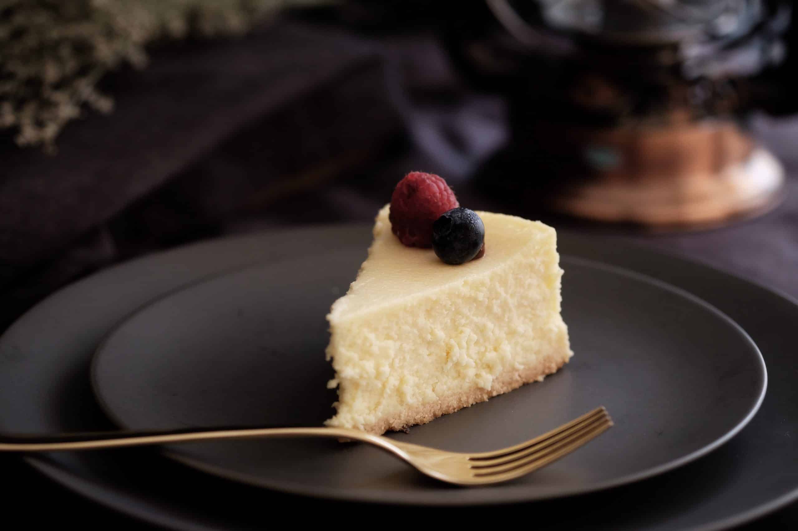 part de cheesecake avec fruits rouges dessus dans une assiette noire avec fourchette dorée à côté